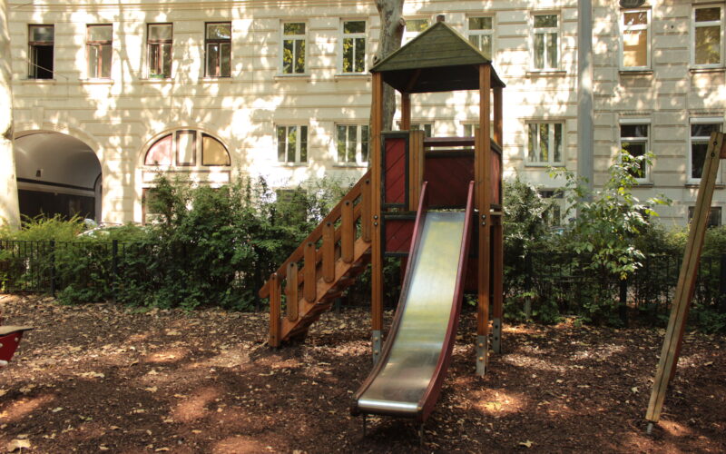 Eine Metallrutsche mit Holztreppe und Unterstand steht in einem schattigen Spielplatzbereich, umgeben von Bäumen und einem weißen Wohnhaus im Hintergrund.