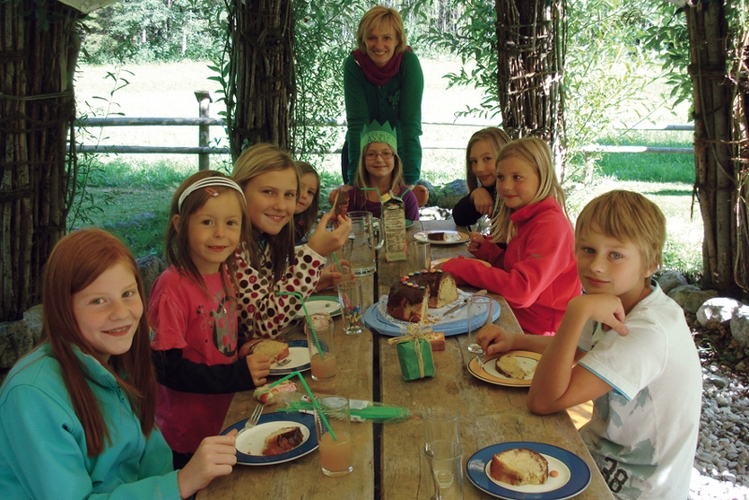 Eine Gruppe von Kindern und ein Erwachsener sitzen lächelnd an einem Holztisch im Freien und essen Kuchen, umgeben von Grün und natürlichem Licht.