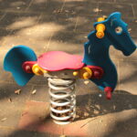 Ein blauer und roter Springreiter in Form eines Pferdes ist auf einem Spielplatz mit einem gefliesten Gummibelag installiert.