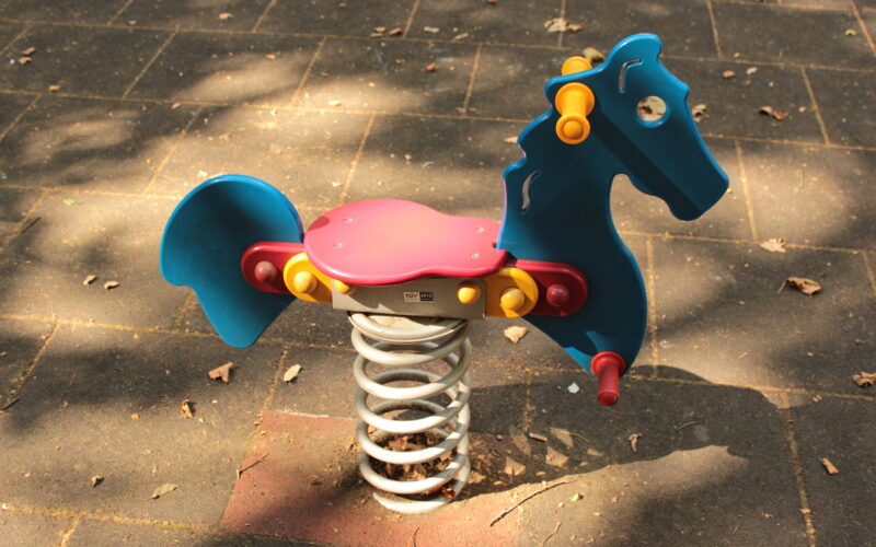 Ein blauer und roter Springreiter in Form eines Pferdes ist auf einem Spielplatz mit einem gefliesten Gummibelag installiert.