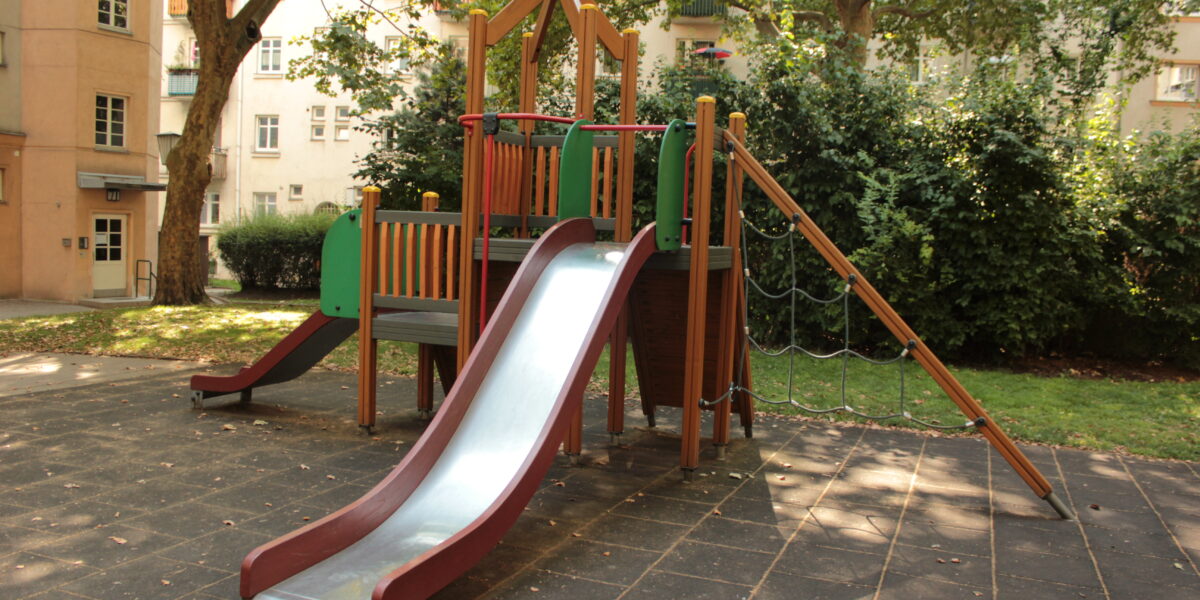 Eine moderne Spielplatzstruktur mit einer Metallrutsche, Kletterseilen und Plattformen auf einer gummierten Oberfläche in einem schattigen Stadtpark.