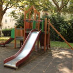 Eine moderne Spielplatzstruktur mit einer Metallrutsche, Kletterseilen und Plattformen auf einer gummierten Oberfläche in einem schattigen Stadtpark.