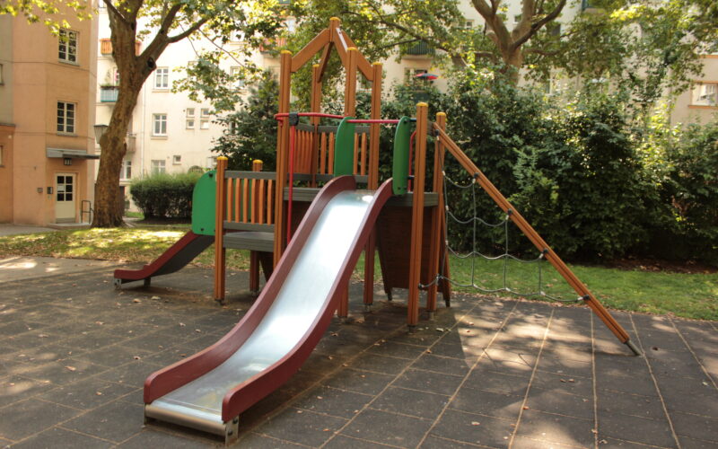 Eine moderne Spielplatzstruktur mit einer Metallrutsche, Kletterseilen und Plattformen auf einer gummierten Oberfläche in einem schattigen Stadtpark.