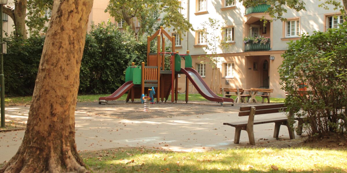 Ein kleiner Spielplatz mit Rutschen und Klettergeräten ist von Bäumen und Bänken in einem Wohnhof umgeben.