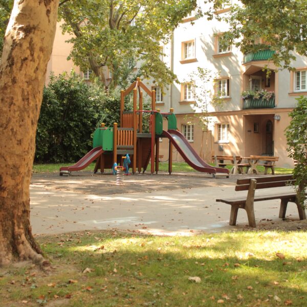 Ein kleiner Spielplatz mit Rutschen und Klettergeräten ist von Bäumen und Bänken in einem Wohnhof umgeben.