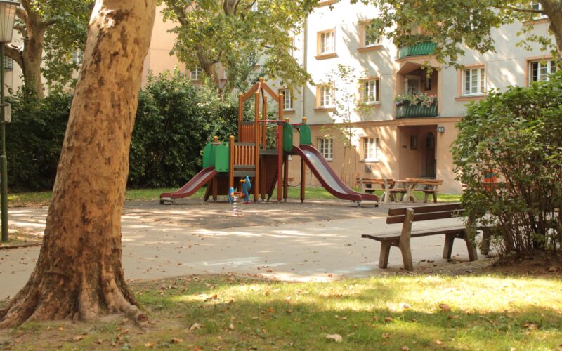 Ein kleiner Spielplatz mit Rutschen und Klettergeräten ist von Bäumen und Bänken in einem Wohnhof umgeben.