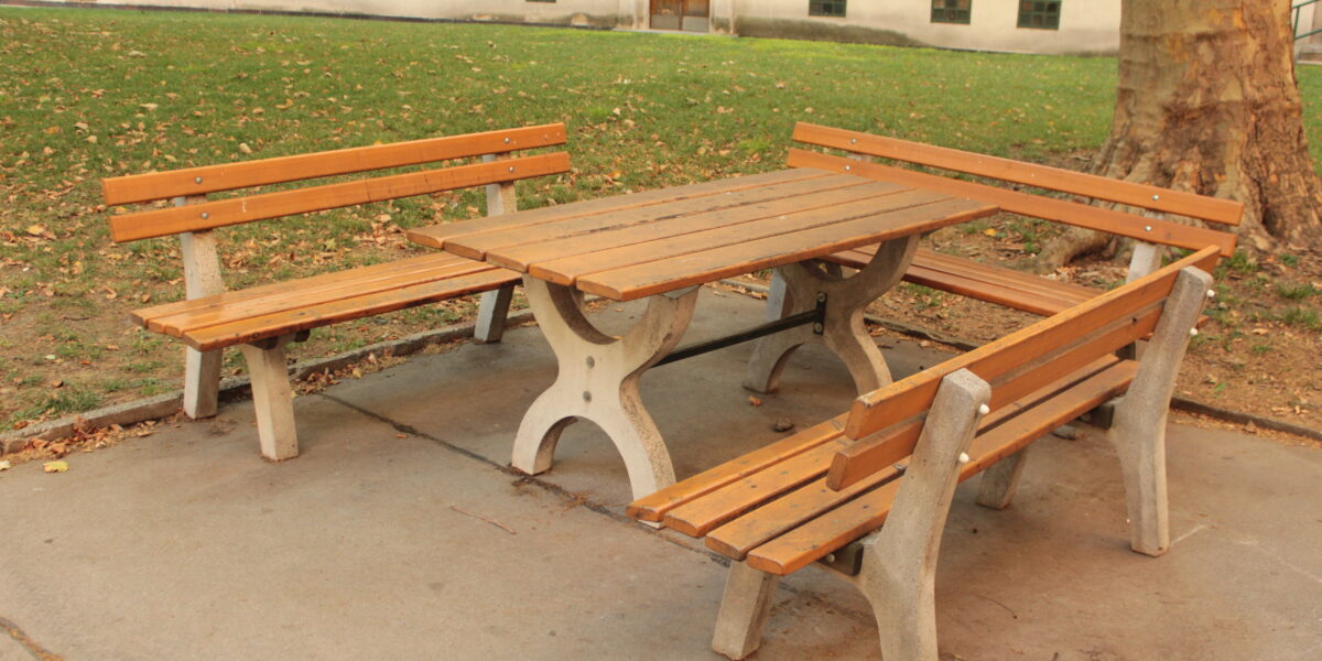 Picknicktisch aus Holz mit zwei Bänken und einer Sitzgelegenheit, aufgestellt auf einer Betonfläche neben einem Baum in einem Park im Freien.