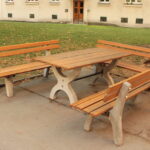 Picknicktisch aus Holz mit zwei Bänken und einer Sitzgelegenheit, aufgestellt auf einer Betonfläche neben einem Baum in einem Park im Freien.