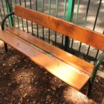 Eine hölzerne Parkbank mit Metallarmlehnen steht auf einer unbefestigten Fläche in der Nähe eines grünen Metallzauns. Auf der Rückenlehne der Bank befindet sich ein Graffiti. Auf dem Boden liegen verstreute Blätter.