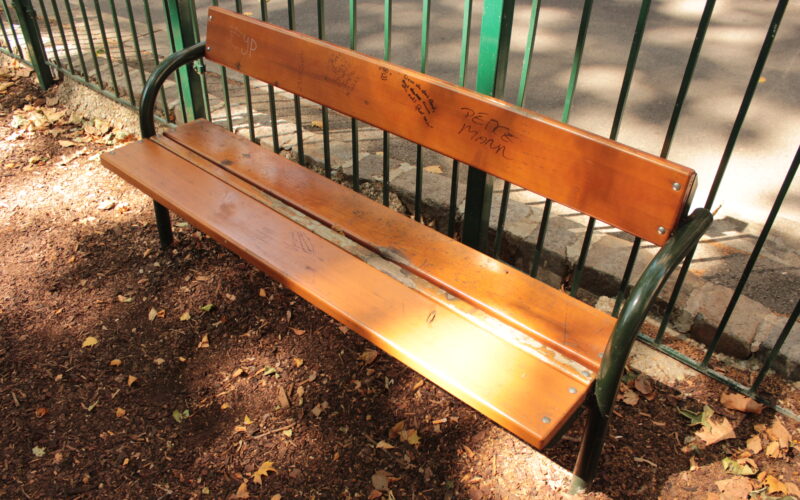 Eine hölzerne Parkbank mit Metallarmlehnen steht auf einer unbefestigten Fläche in der Nähe eines grünen Metallzauns. Auf der Rückenlehne der Bank befindet sich ein Graffiti. Auf dem Boden liegen verstreute Blätter.