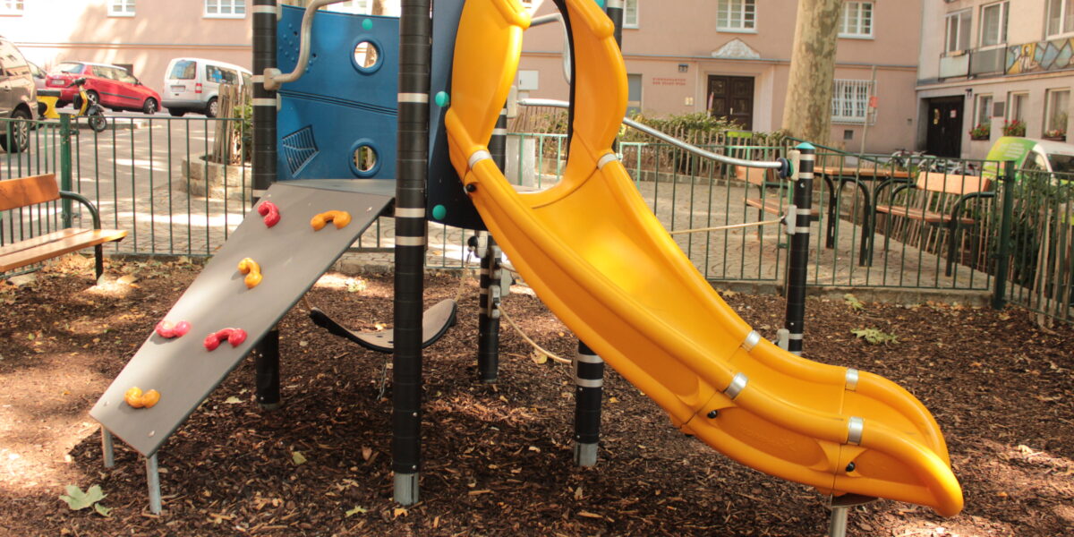 Ein Spielplatz mit einer gelben Rutsche, einer kleinen Kletterwand und einer blauen Platte, umgeben von Mulch und einem Metallzaun in einem städtischen Wohngebiet.