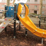 Ein Spielplatz mit einer gelben Rutsche, einer kleinen Kletterwand und einer blauen Platte, umgeben von Mulch und einem Metallzaun in einem städtischen Wohngebiet.