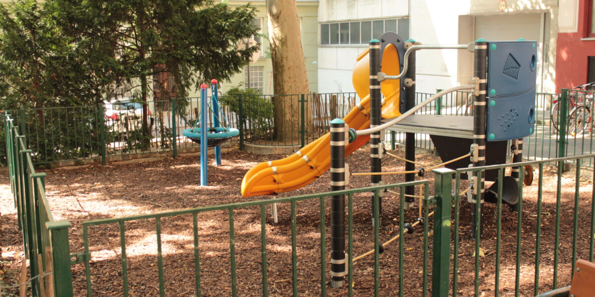 Ein kleiner eingezäunter Spielplatz mit einer gelben Rutsche, einem Klettergerüst und drehenden Spielgeräten, umgeben von Bäumen und Gebäuden.