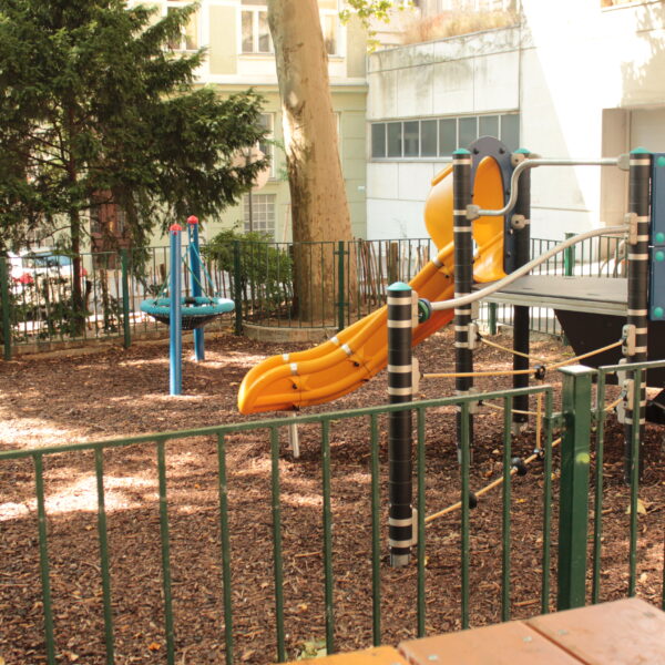 Ein kleiner eingezäunter Spielplatz mit einer gelben Rutsche, einem Klettergerüst und drehenden Spielgeräten, umgeben von Bäumen und Gebäuden.