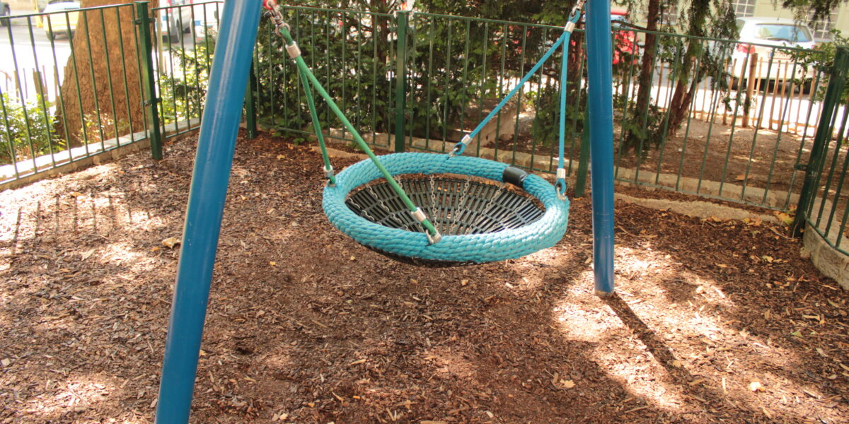 Eine runde Netzschaukel mit blauem Rahmen steht auf einem eingezäunten Spielplatz, umgeben von Mulch und Bäumen, wobei das Sonnenlicht Schatten auf den Boden wirft.
