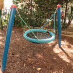 Eine runde Netzschaukel mit blauem Rahmen steht auf einem eingezäunten Spielplatz, umgeben von Mulch und Bäumen, wobei das Sonnenlicht Schatten auf den Boden wirft.