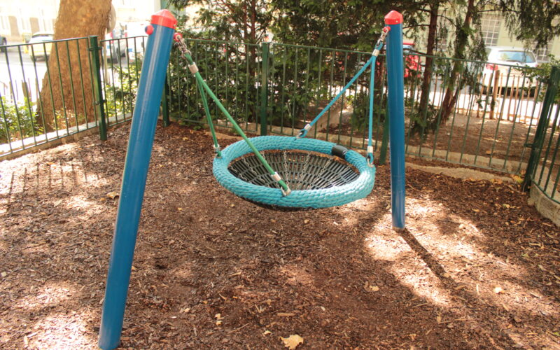 Eine runde Netzschaukel mit blauem Rahmen steht auf einem eingezäunten Spielplatz, umgeben von Mulch und Bäumen, wobei das Sonnenlicht Schatten auf den Boden wirft.