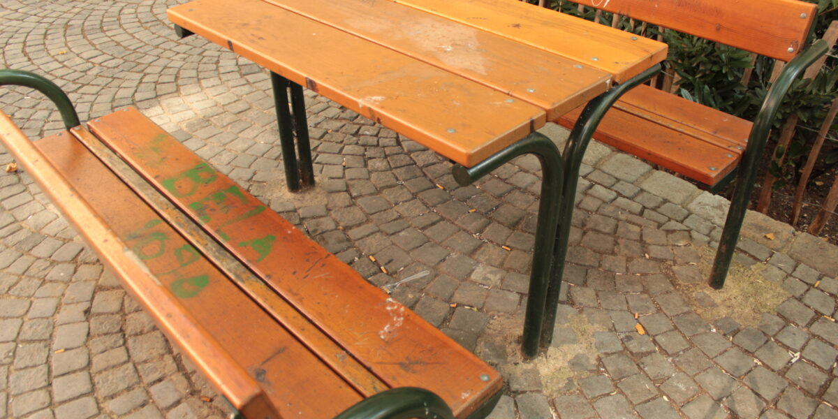 Ein Holztisch für den Außenbereich mit zwei Bänken auf einer Kopfsteinpflasterfläche. Die Bank und der Tisch haben grüne Metallbeine und einige Graffiti auf dem Holz.