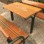 Ein Holztisch für den Außenbereich mit zwei Bänken auf einer Kopfsteinpflasterfläche. Die Bank und der Tisch haben grüne Metallbeine und einige Graffiti auf dem Holz.