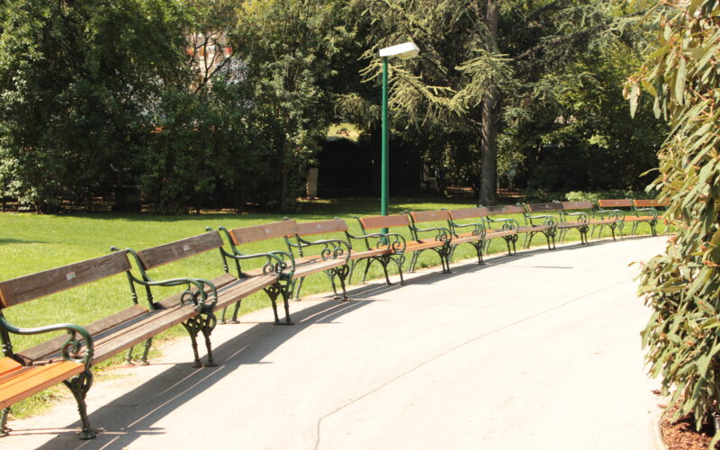 Eine geschwungene Reihe leerer Holzbänke säumt einen gepflasterten Weg in einem sonnendurchfluteten Park, umgeben von grünen Bäumen und Büschen.