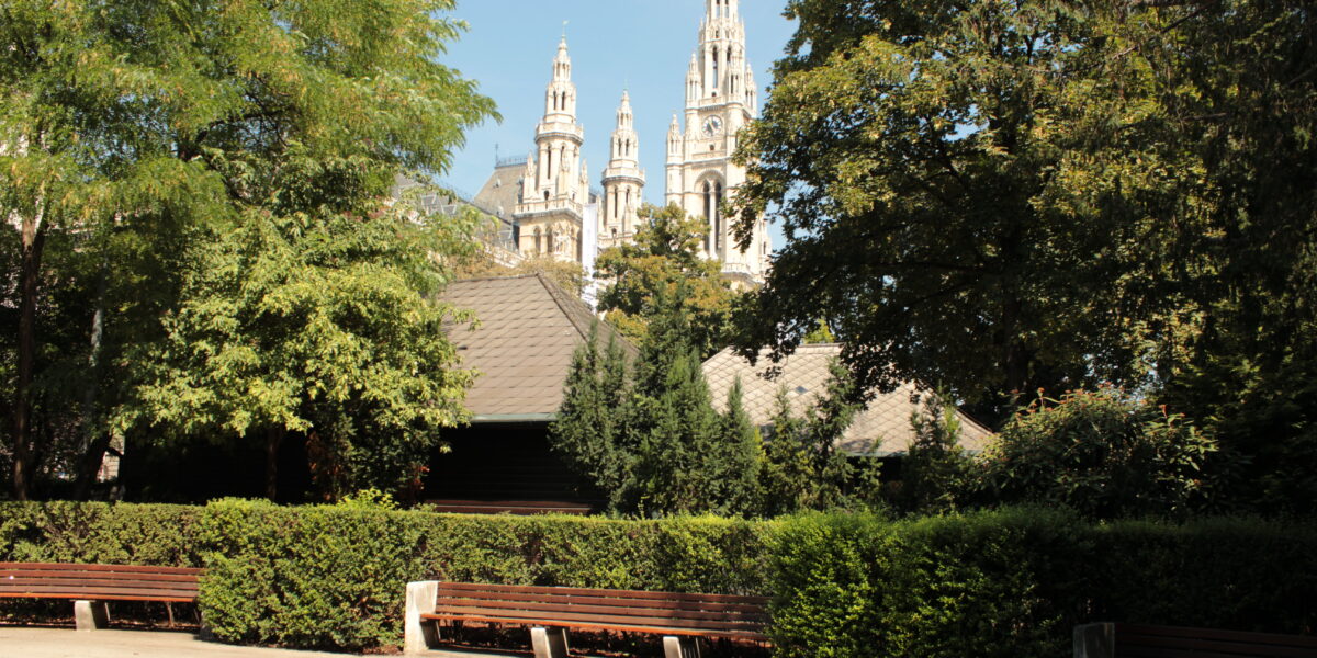 Ein Park mit leeren Bänken, grünen Bäumen und den Türmen eines großen historischen Gebäudes, die im Hintergrund unter einem blauen Himmel sichtbar sind.