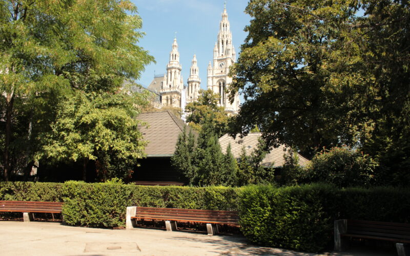 Ein Park mit leeren Bänken, grünen Bäumen und den Türmen eines großen historischen Gebäudes, die im Hintergrund unter einem blauen Himmel sichtbar sind.