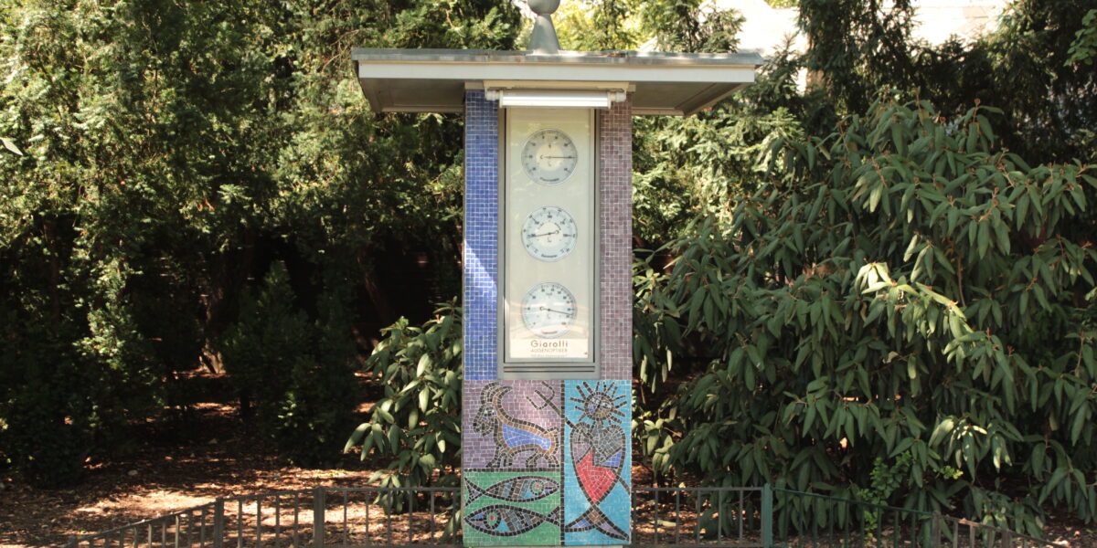 Eine freistehende Uhrensäule im Freien zeigt drei Uhren über einem farbenfrohen Mosaik, umgeben von Grün auf einer gepflasterten Fläche.