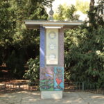 Eine freistehende Uhrensäule im Freien zeigt drei Uhren über einem farbenfrohen Mosaik, umgeben von Grün auf einer gepflasterten Fläche.