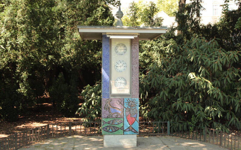 Eine freistehende Uhrensäule im Freien zeigt drei Uhren über einem farbenfrohen Mosaik, umgeben von Grün auf einer gepflasterten Fläche.