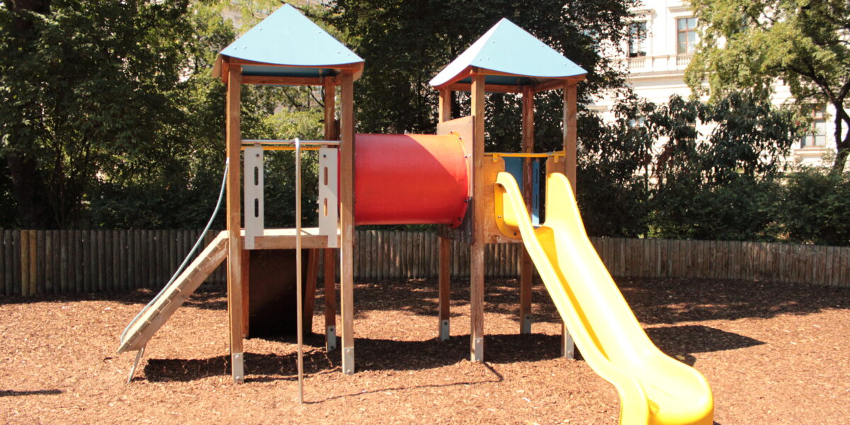 Spielplatz im Freien mit zwei Türmen, die durch einen roten Tunnel verbunden sind, einer Metallrutsche auf der linken Seite und einer gelben Kunststoffrutsche auf der rechten Seite, umgeben von Mulch und Bäumen.