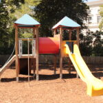Spielplatz im Freien mit zwei Türmen, die durch einen roten Tunnel verbunden sind, einer Metallrutsche auf der linken Seite und einer gelben Kunststoffrutsche auf der rechten Seite, umgeben von Mulch und Bäumen.