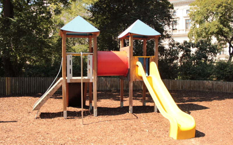 Spielplatz im Freien mit zwei Türmen, die durch einen roten Tunnel verbunden sind, einer Metallrutsche auf der linken Seite und einer gelben Kunststoffrutsche auf der rechten Seite, umgeben von Mulch und Bäumen.