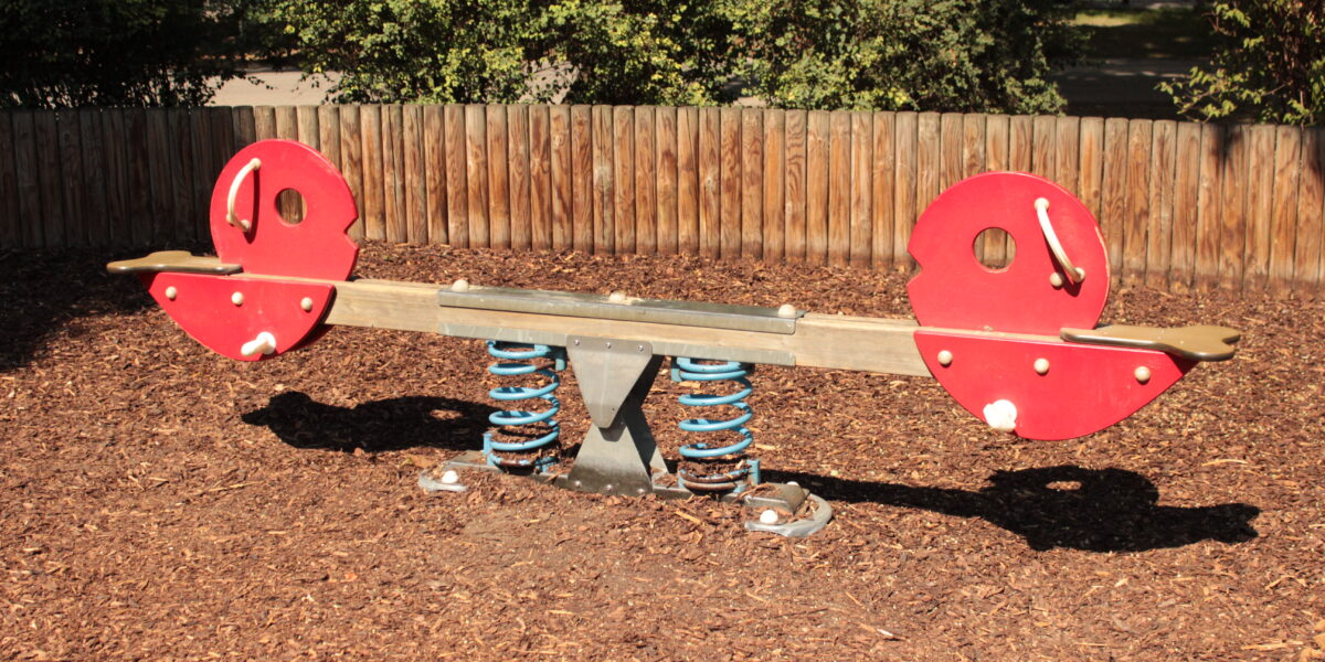 Eine rote und braune gefederte Wippe mit Metallgriffen steht auf Holzspänen auf einem Spielplatz, mit einem Holzzaun und Grünpflanzen im Hintergrund.