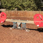 Eine rote und braune gefederte Wippe mit Metallgriffen steht auf Holzspänen auf einem Spielplatz, mit einem Holzzaun und Grünpflanzen im Hintergrund.