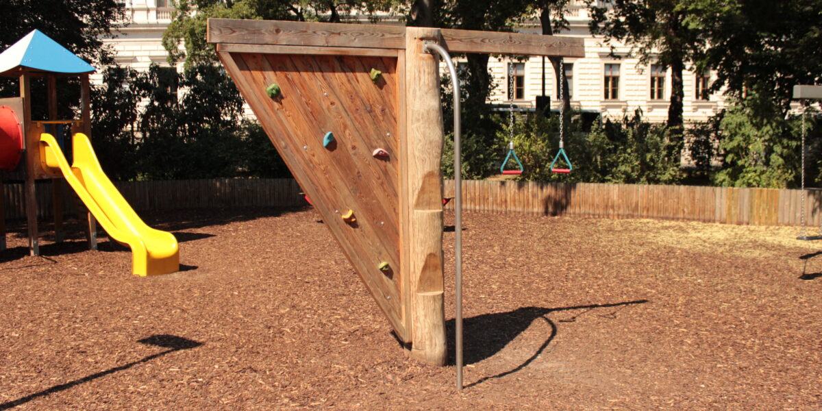 Auf einem Spielplatz mit Mulchboden steht eine dreieckige Kletterwand aus Holz mit Griffen und einer senkrechten Metallstange.
