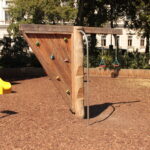 Auf einem Spielplatz mit Mulchboden steht eine dreieckige Kletterwand aus Holz mit Griffen und einer senkrechten Metallstange.