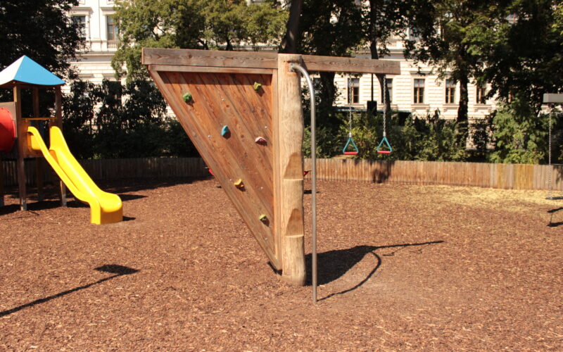 Auf einem Spielplatz mit Mulchboden steht eine dreieckige Kletterwand aus Holz mit Griffen und einer senkrechten Metallstange.