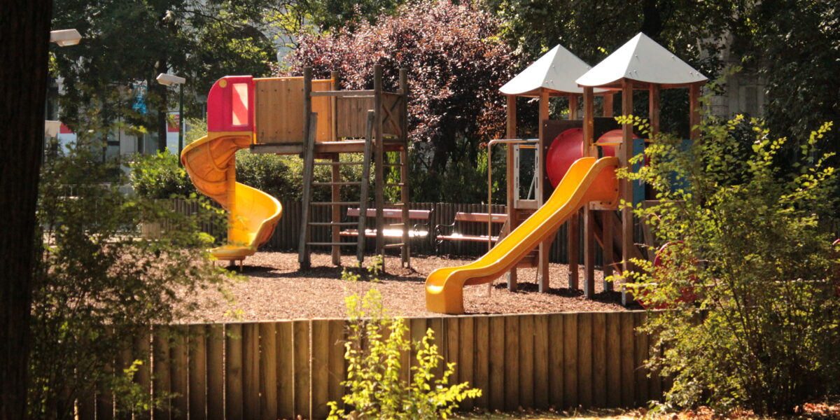 Ein Spielplatz mit Rutschen, Klettergerüsten und Bänken, umgeben von Bäumen und einem Holzzaun an einem sonnigen Tag.