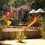 Ein Spielplatz mit Rutschen, Klettergerüsten und Bänken, umgeben von Bäumen und einem Holzzaun an einem sonnigen Tag.