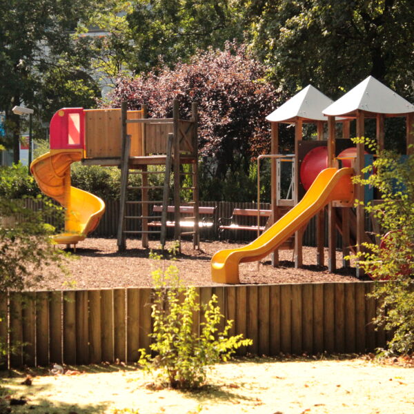 Ein Spielplatz mit Rutschen, Klettergerüsten und Bänken, umgeben von Bäumen und einem Holzzaun an einem sonnigen Tag.