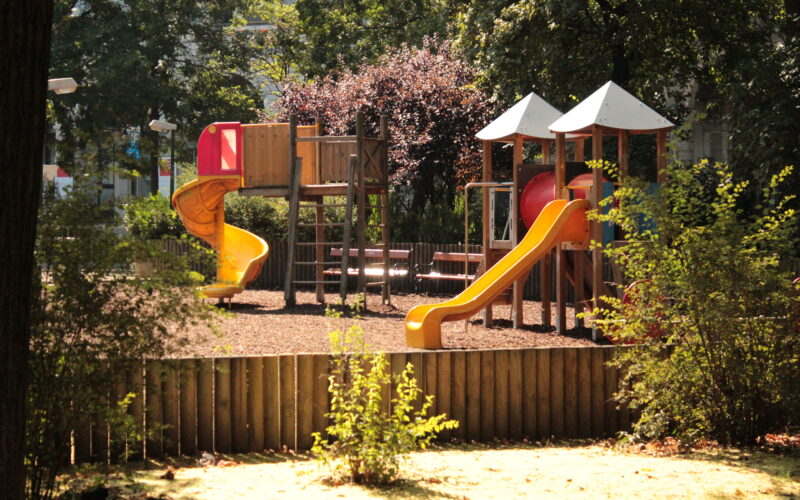 Ein Spielplatz mit Rutschen, Klettergerüsten und Bänken, umgeben von Bäumen und einem Holzzaun an einem sonnigen Tag.