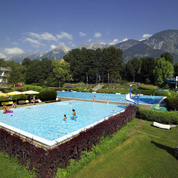 Öffentliches Freibad mit schwimmenden Menschen, umgeben von grünem Gras, Bäumen, Liegestühlen, Wasserrutschen und Bergen im Hintergrund unter einem klaren Himmel.