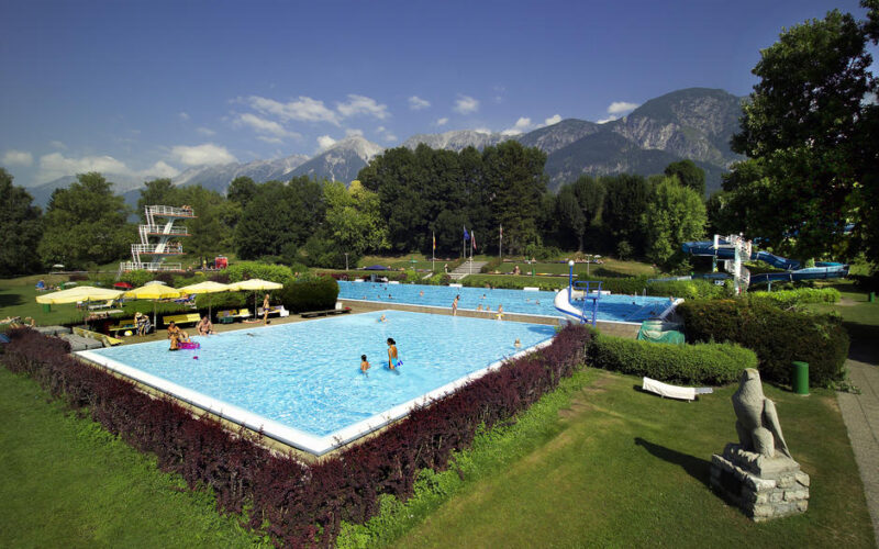 Öffentliches Freibad mit schwimmenden Menschen, umgeben von grünem Gras, Bäumen, Liegestühlen, Wasserrutschen und Bergen im Hintergrund unter einem klaren Himmel.
