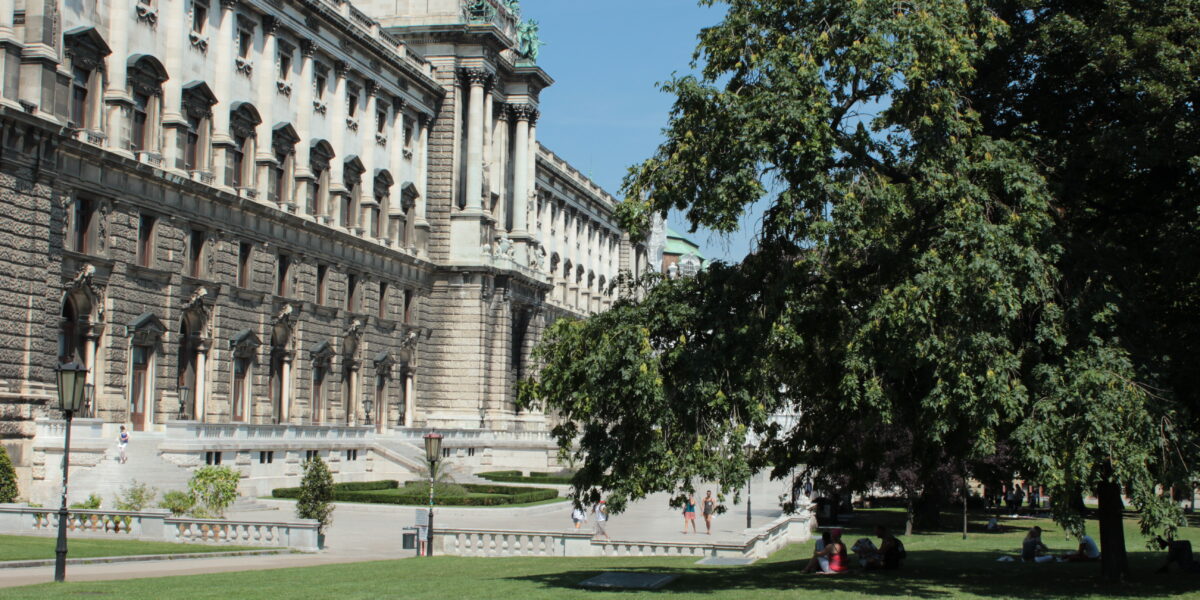 Ein großes historisches Gebäude mit kunstvoller Architektur steht neben einem grünen Rasen und Bäumen, und Menschen gehen unter einem klaren Himmel im Park spazieren und sitzen.