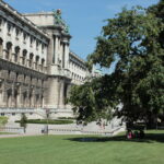 Ein großes historisches Gebäude mit kunstvoller Architektur steht neben einem grünen Rasen und Bäumen, und Menschen gehen unter einem klaren Himmel im Park spazieren und sitzen.