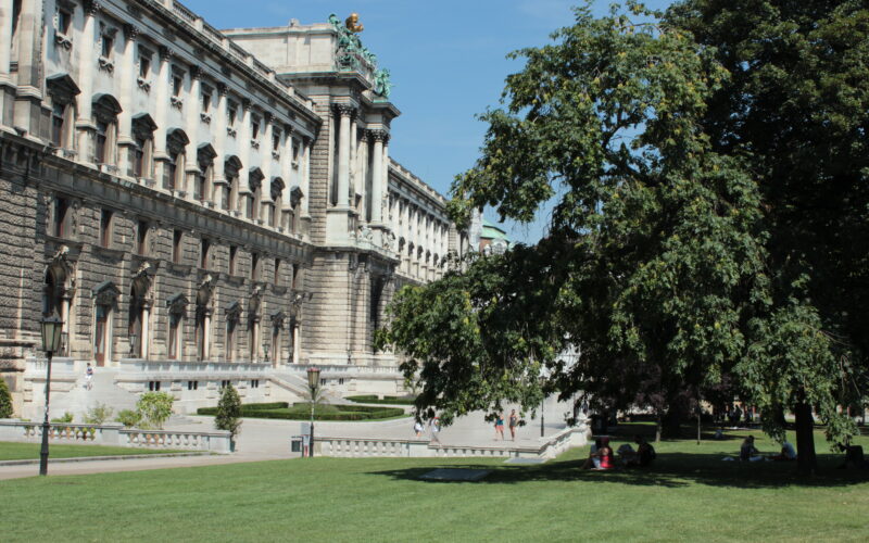 Ein großes historisches Gebäude mit kunstvoller Architektur steht neben einem grünen Rasen und Bäumen, und Menschen gehen unter einem klaren Himmel im Park spazieren und sitzen.