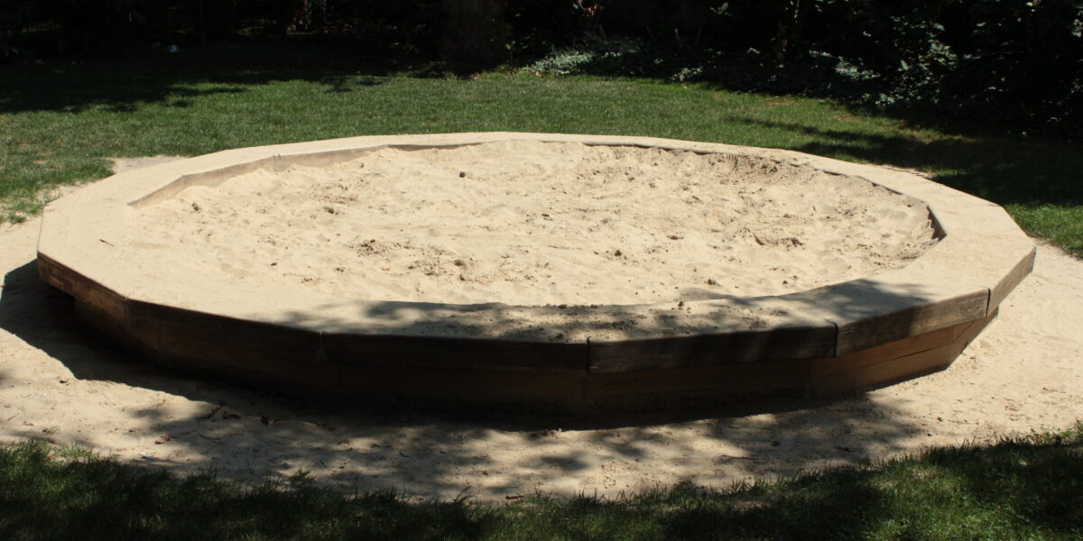 Ein leerer achteckiger Sandkasten mit Holzseiten steht auf Gras in einem schattigen Außenbereich.
