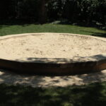 Ein leerer achteckiger Sandkasten mit Holzseiten steht auf Gras in einem schattigen Außenbereich.