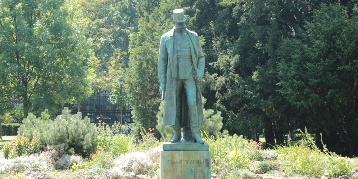 Eine Bronzestatue eines Mannes in langem Mantel und Hut steht auf einem Steinsockel, umgeben von Grün in einem Park.