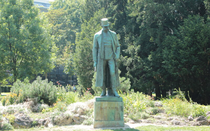 Eine Bronzestatue eines Mannes in langem Mantel und Hut steht auf einem Steinsockel, umgeben von Grün in einem Park.